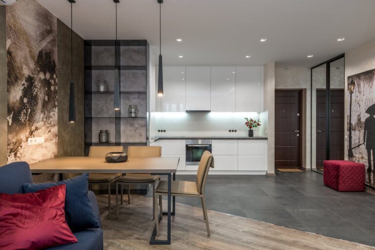 Wooden dining table with chairs separating living room with sofa from kitchen zone with minimalist white furniture and modern appliances in spacious studio apartment