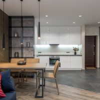 Wooden dining table with chairs separating living room with sofa from kitchen zone with minimalist white furniture and modern appliances in spacious studio apartment